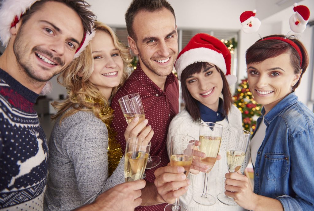 fem unge mennesker, der fejrer julen med et glas champagne i hånden og med festlige julehatte på.